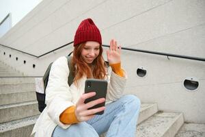 Happy college girl talks with friends on video chat smartphone app, sits on stairs outdoors uses her mobile phone application, waves hand at telephone camera photo