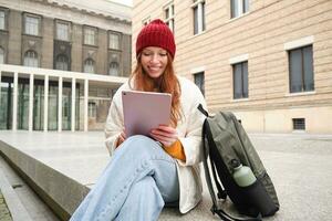 Redhead girl smiles, sits outdoors near building with digital tablet, thermos and backpack, connects to public internet and searches smth online on her gadget photo