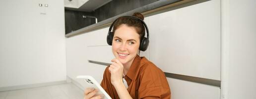 Positive young woman, sits at home with wireless headphones and smartphone, smiles and looks happy photo