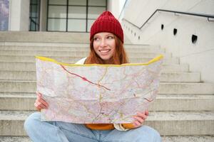 joven sonriente pelirrojo chica, turista se sienta en escalera al aire libre con ciudad papel mapa, mirando para dirección, viajero mochilero explora ciudad y mira para Turismo foto