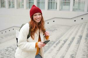 Smiling redhead girl, student tourist walks around city, goes up the stairs, looks at mobile phone map to get around town, sends message on smartphone photo