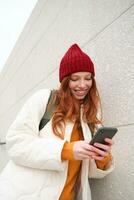alegre niña turista mira a móvil teléfono, textos mensaje en teléfono inteligente social medios de comunicación solicitud, camina alrededor ciudad, mira para Turismo en móvil aplicación foto