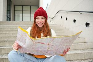 hermosa niña turista se sienta en escalera con ciudad mapa, planes su viaje, mira para dirección mientras de viaje alrededor ciudad, búsquedas ruta para Turismo foto