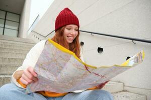 hermosa niña turista se sienta en escalera con ciudad mapa, planes su viaje, mira para dirección mientras de viaje alrededor ciudad, búsquedas ruta para Turismo foto