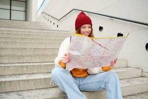 hermosa niña turista se sienta en escalera con ciudad mapa, planes su viaje, mira para dirección mientras de viaje alrededor ciudad, búsquedas ruta para Turismo foto