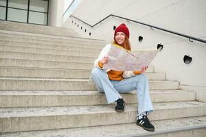 Beautiful girl tourist sits on stairs with city map, plans her journey, looks for direction while travelling around town, searches route for sightseeing photo