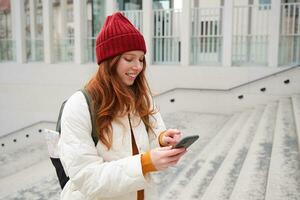 Smiling redhead girl, student tourist walks around city, goes up the stairs, looks at mobile phone map to get around town, sends message on smartphone photo