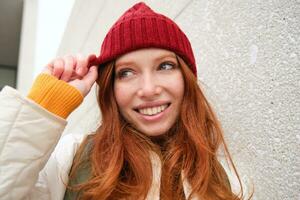 Stylish redhead girl in red hat, smiles and looks happy, poses outdoors on street, looks relaxed and lively photo