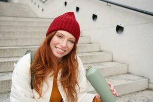 Happy redhead woman, tourist with thermos, drinks her hot tea, coffee from travel flask, restests during her travelling in city and sightseeing photo