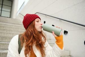 Smiling traveler, redhead girl tourist sits on stairs with flask, drinks hot coffee from thermos while travelling and sightseeing around foreign city, sits on stairs and rests photo