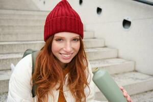 Smiling traveler, redhead girl tourist sits on stairs with flask, drinks hot coffee from thermos while travelling and sightseeing around foreign city, sits on stairs and rests photo