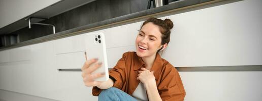 Portrait of beautiful smiling woman, talking to friend on smartphone video chat, connects to online conversation on mobile app, sitting on kitchen floor photo
