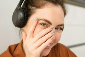 Cute smiling girl, blushes, hides face behind hand and laughing, wears wireless headphones photo