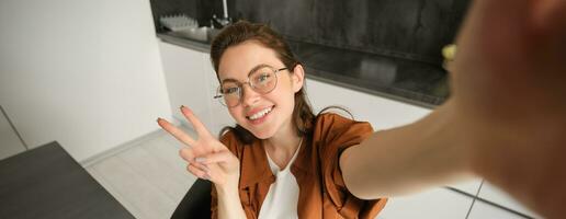 Selfie of cute young woman in glasses, posing at home in kitchen, taking pictures on her mobile phone, smiling and showing peace sign photo