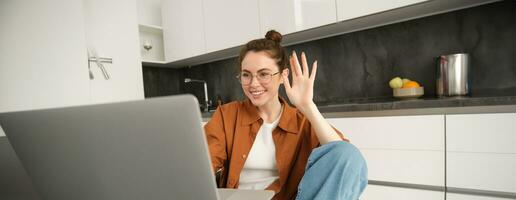 Cheerful young woman in glasses, saying hello, waving hand at laptop, talking to friend online, video chatting, sitting in kitchen with computer photo