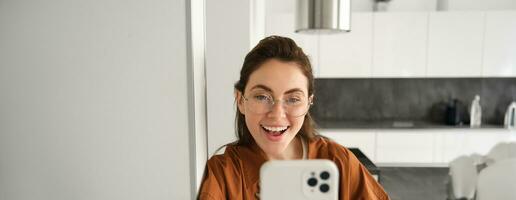 cerca arriba retrato de contento niña toma selfie en teléfono inteligente, posando con móvil teléfono, mirando emocionado y asombrado, sonriente a teléfono cámara, utilizando social medios de comunicación aplicación foto