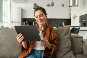 retrato de joven mujer sentado en sofá en vivo habitación, acecho películas en digital tableta, leyendo en dispositivo, disfrutando fin de semana a hogar foto