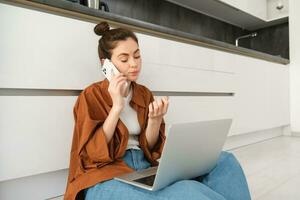 mujer mirando aburrido y aburrido mientras sentado en piso y escuchando a conversación, tiene ordenador portátil en vueltas, esperando en línea, haciendo teléfono llamada foto