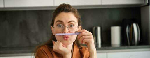 Close up portrait of funny, silly young woman, playing with pen, holding pencil on top of her lip and grimacing, sitting in kitchen photo