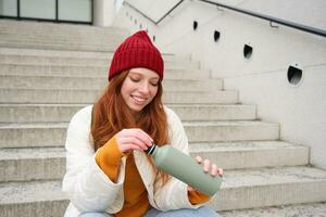 Smiling traveler, redhead girl tourist sits on stairs with flask, drinks hot coffee from thermos while travelling and sightseeing around foreign city, sits on stairs and rests photo