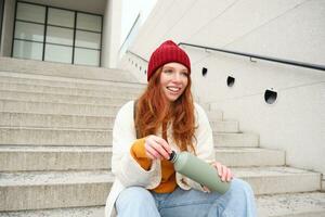 Smiling traveler, redhead girl tourist sits on stairs with flask, drinks hot coffee from thermos while travelling and sightseeing around foreign city, sits on stairs and rests photo
