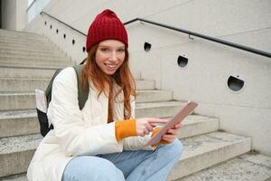 Young stylish girl, redhead female students sits on stairs outdoors with digital tablet, reads, uses social media app on gadget, plays games while waits on street photo