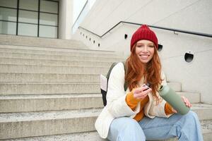 Happy redhead woman, tourist with thermos, drinks her hot tea, coffee from travel flask, restests during her travelling in city and sightseeing photo