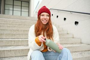 Smiling traveler, redhead girl tourist sits on stairs with flask, drinks hot coffee from thermos while travelling and sightseeing around foreign city, sits on stairs and rests photo