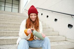 Happy redhead woman, tourist with thermos, drinks her hot tea, coffee from travel flask, restests during her travelling in city and sightseeing photo