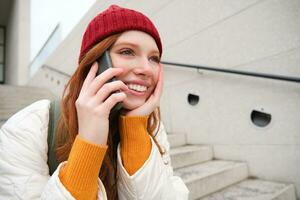 Beautiful smiling redhead female model, sits on street and talks on mobile phone, uses smartphone app to call abroad, laughing during telephone conversation photo