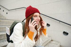 Beautiful smiling redhead female model, sits on street and talks on mobile phone, uses smartphone app to call abroad, laughing during telephone conversation photo