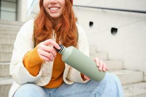 Smiling traveler, redhead girl tourist sits on stairs with flask, drinks hot coffee from thermos while travelling and sightseeing around foreign city, sits on stairs and rests photo