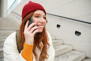 joven elegante pelirrojo niña en rojo sombrero, se sienta en calle y negociaciones en móvil teléfono, tiene teléfono conversación, anillos su amigo mientras relaja al aire libre foto