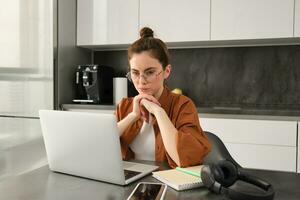 Freelancing, work from home concept. Young woman sitting with laptop in kitchen, freelancer digital nomad doing her job on remote, using computer, looking thoughtful, thinking photo