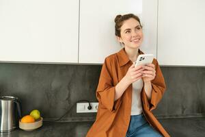 Image of young woman with smartphone, looking with thoughtful expression, buying online using mobile app, orders food delivery with phone application photo
