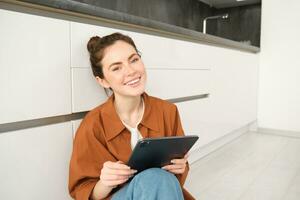 Portrait of young beautiful woman sitting on kitchen floor with digital tablet, browsing news feed, social media app on gadget, smiling and looking happy photo