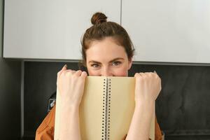 Portrait of smiling woman hiding her smile behind notebook, giggling and looking at camera flirty, standing in the kitchen with notes photo