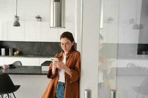 Portrait of cute young woman standing at home with smartphone, reading message on mobile phone, using application, order online, paying utility bill in app photo