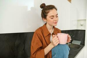 retrato de hermosa joven mujer disfrutando su Mañana taza de aromático café, Bebiendo té mientras sentado en cocina y contemplando soleado día foto