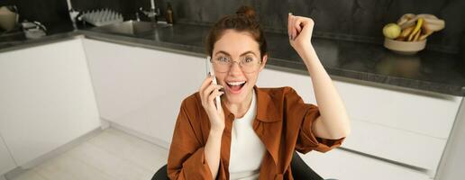 Portrait of woman receiving great news over the phone. Girl talking on mobile telephone and celebrating, laughing and making fist pump, dancing on her chair in kitchen photo