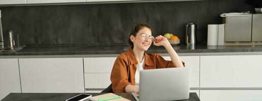 Portrait of young woman, business owner in glasses, sitting with laptop in kitchen, working from home. Student studying, doing homework on computer photo