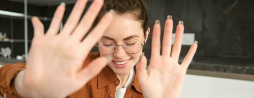 Portrait of cute and romantic young woman in glasses, covers her face, extends hands to block camera, asks to stop taking pictures photo