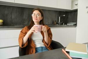 Portrait of young professional, girl freelancer sitting in kitchen and enjoying cup of coffee, sitting with documents, taking break from studying and smiling, looking outside window photo