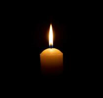 A single burning candle flame or light glowing on a big yellow candle on black or dark background on table in church for Christmas, funeral or memorial service with copy space photo