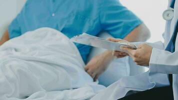 Senior doctor and young male patient who lie on the bed while checking pulse, consult and explain with nurse taking note and supporting in hospital wards. video