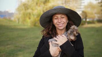 bellissimo sorridente contento donna nel un' grande grigio cappello ritratto nel il parco Tenere un' piccolo yorkie cane nel sua braccia e baci lui 4k video