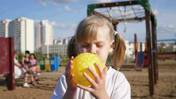 poco ragazza bambino ritratto si gonfia un' giallo Palloncino su il terreno di gioco nel estate 4k lento movimento video