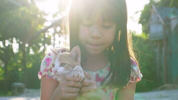 carino ragazza con rosso gattino. un' piccolo gattino nel il braccia di un' poco ragazza. un' bambino detiene un' rosso gatto con tenerezza e amore e sorrisi felicemente nel il Giardino dietro la casa all'aperto nel il luce del sole. video