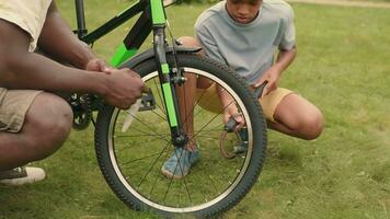 African dad helps his son repair his bike on the lawn near the house. video