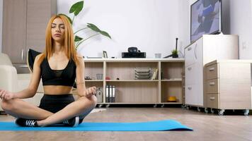 femme pratiquant yoga sur une bleu tapis dans vivant pièce tandis que le la télé dans sur dans le Contexte. elle est une magnifique arabe mixte raxe l'ethnie Jeune femme dans lotus pose video